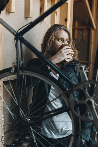 Stylish young man with long hair standing near hipster bicycle and drinking coffee