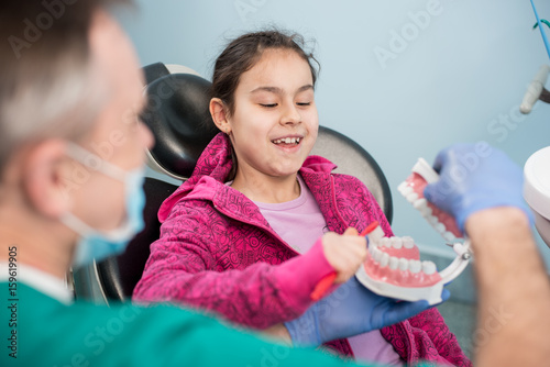 Photography Smiling girl in dentist chair educating about proper tooth-brushing by her pediatric dentist, using dental jaw model and toothbrush in dental clinic