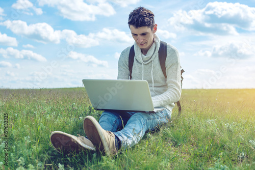 Young man sitting on a green meadow with laptop wireless on the background of blue cloudy sky