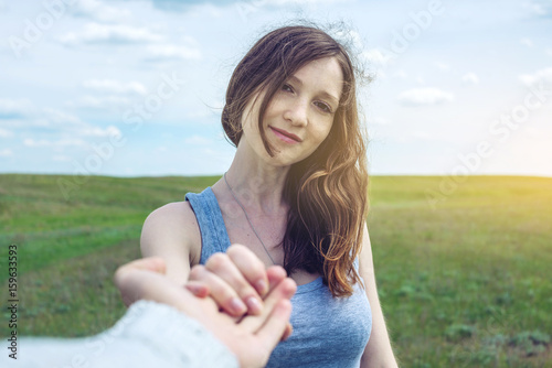 Follow me, Attractive brunette girl holding the hand of the leads in a clean green field, steppe with clouds