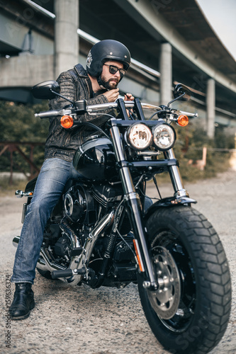Young brutal man in a black jacket and glasses on motorcycle. Male biker wears a black helmet.