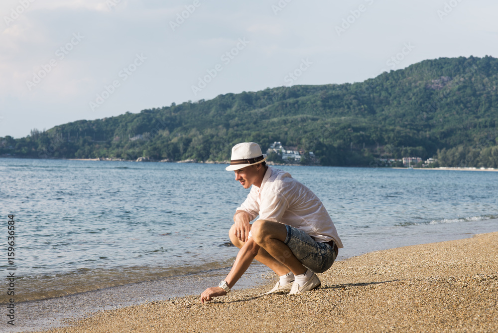 young handsome man in white shirt and hat sit down on his knees by beautiful beach and looking for seashells in the sand. outdoor fashion lifestyle image