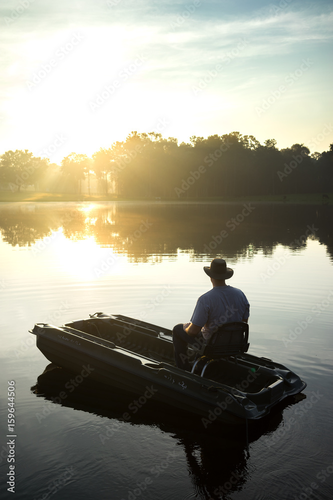 Man in small fishing boat on lake river water at sunrise sunset dawn early morning dusk with sun rays and trees on horizon feeling peaceful relaxed serene calm meditative alone sad lonely on vacation