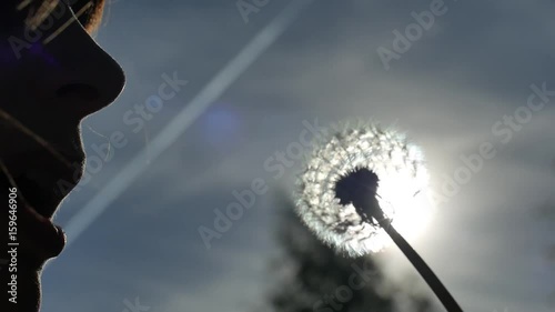 A girl blowing on a dandelion parashutiki fly out in the wind. The bright sun shines with backlight. Concept of summer fun freedom of youth. photo