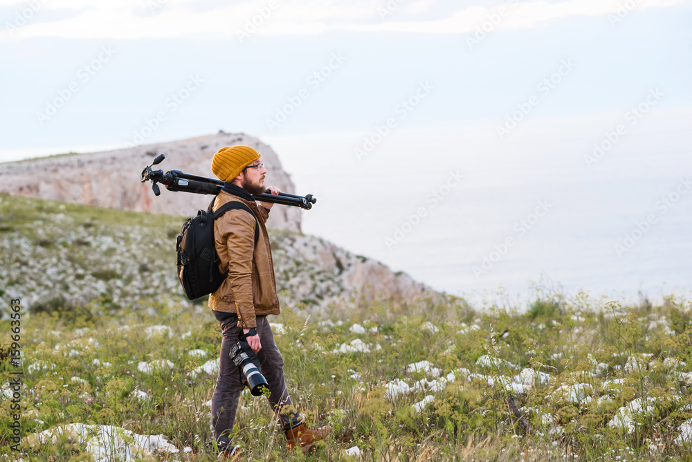 Obraz premium Young photographer standing on a cliff with tripod on his shoulder and camera in hand