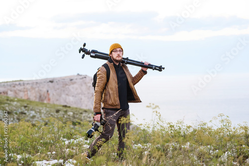 Young photographer walking in the nature with tripod on his shoulder and camera in hand