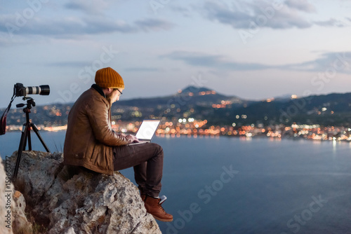 Young attractive photographer sitting on rock and working on laptop with view of sea and mountains