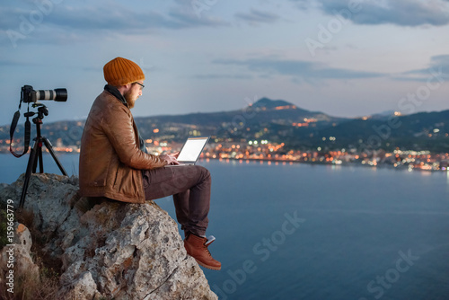 Young attractive photographer sitting on rock and working on laptop with view of sea and mountains © bodiaphoto