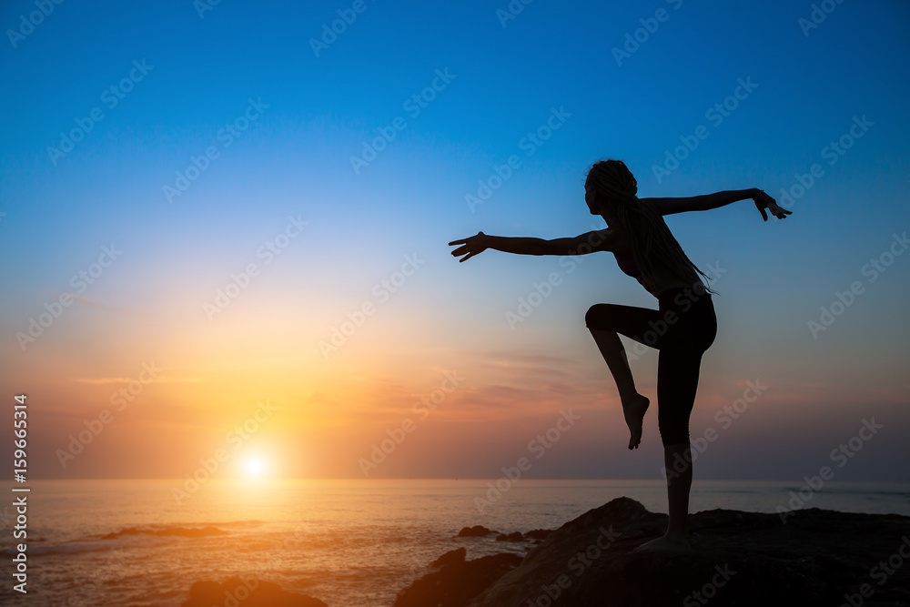 Dancing silhouette of young flexible woman on the sea coast during sunset.
