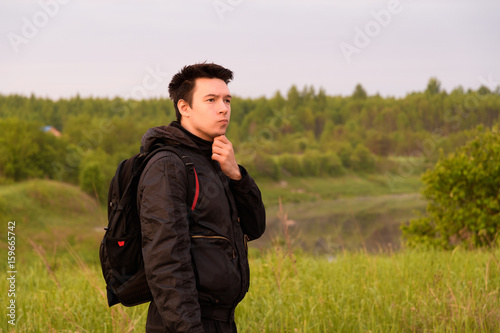 The guy in black clothes with a backpack thoughtfully scratches his chin, on the road to greenery