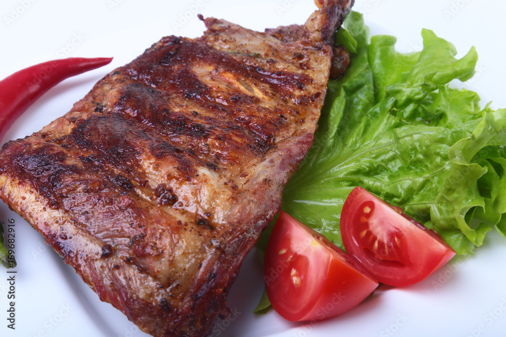 Pork ribs on grill and chili pepper with tomato, lettuce leaves on white plate Pork ribs on grill and chili pepper with tomato, lettuce leaves on white plate
