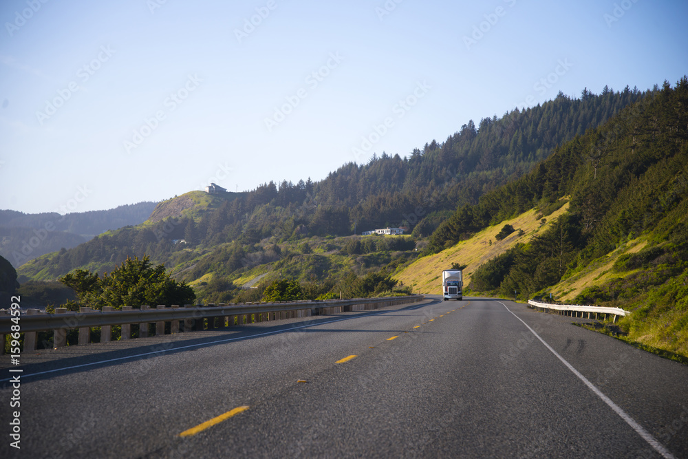 Classic semi truck on spectacular highway with fence and green trees on the hills Classic semi truck on spectacular highway with fence and green trees on the hills
