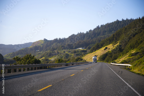 Classic semi truck on spectacular highway with fence and green trees on the hills