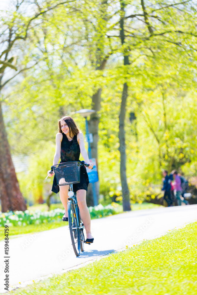 Portrait view of girl on bicycle wearing on black short dress. Young happy Woman riding along road on green spring outdoor Park. Sporty young girl riding a bicycle on a sunny morning, view from face Portrait view of girl on bicycle wearing on black short dress. Young happy Woman riding along road on green spring outdoor Park. Sporty young girl riding a bicycle on a sunny morning, view from face