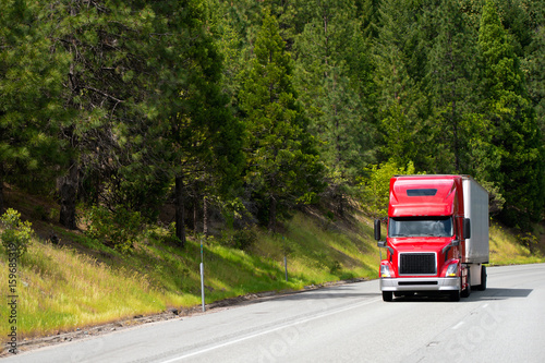 Modern red semi truck with dry van trailer driving on road with green trees forest on the roadside hills