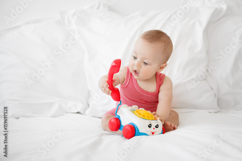 cute baby sitting on a white bed playing with a toy phone