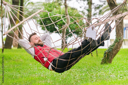 Bearded man in a red vest sleeps in a hammock in the park