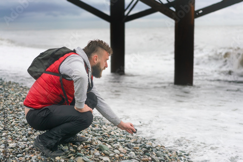 Man in a red vest and backpack sitting near the sea on a pebble beach
