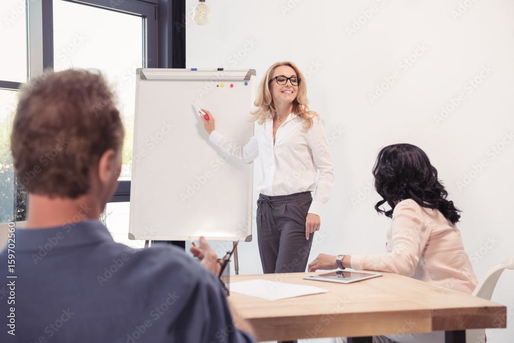 Smiling businesswoman in eyeglasses pointing at blank whiteboard and looking at colleagues during presentation Smiling businesswoman in eyeglasses pointing at blank whiteboard and looking at colleagues during presentation