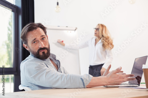 Stressed bearded businessman using laptop while blonde businesswoman working with whiteboard behind