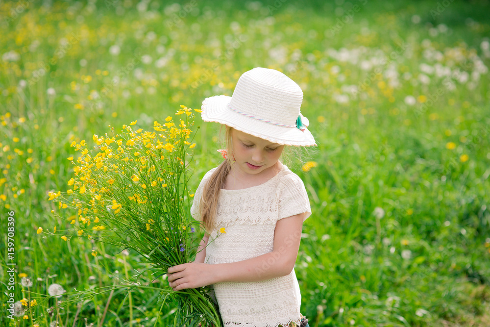 Little beautiful smiling blonde child girl 4-5 years in hat with fields in the field with yellow flowers, with a bouquet in hands