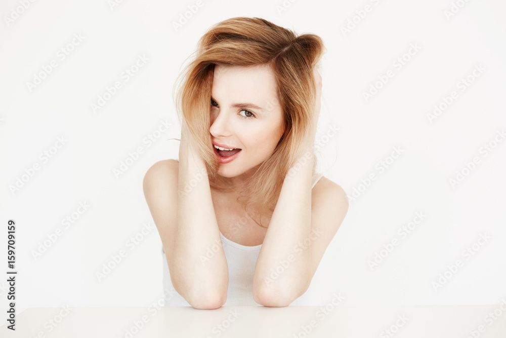 Cheerful natural blonde girl smiling looking at camera sitting at table over white background. Beauty and health concept. Opened mouth. Cheerful natural blonde girl smiling looking at camera sitting at table over white background. Beauty and health concept. Opened mouth.