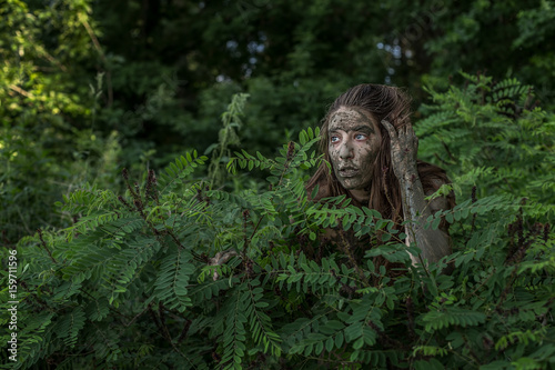 Muddy Amazon brown-haired girl hiding behind a bush in the woods
