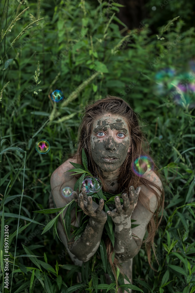 Muddy Amazon brown-haired girl hiding behind a bush in the woods, while soap bubbles flying around her
