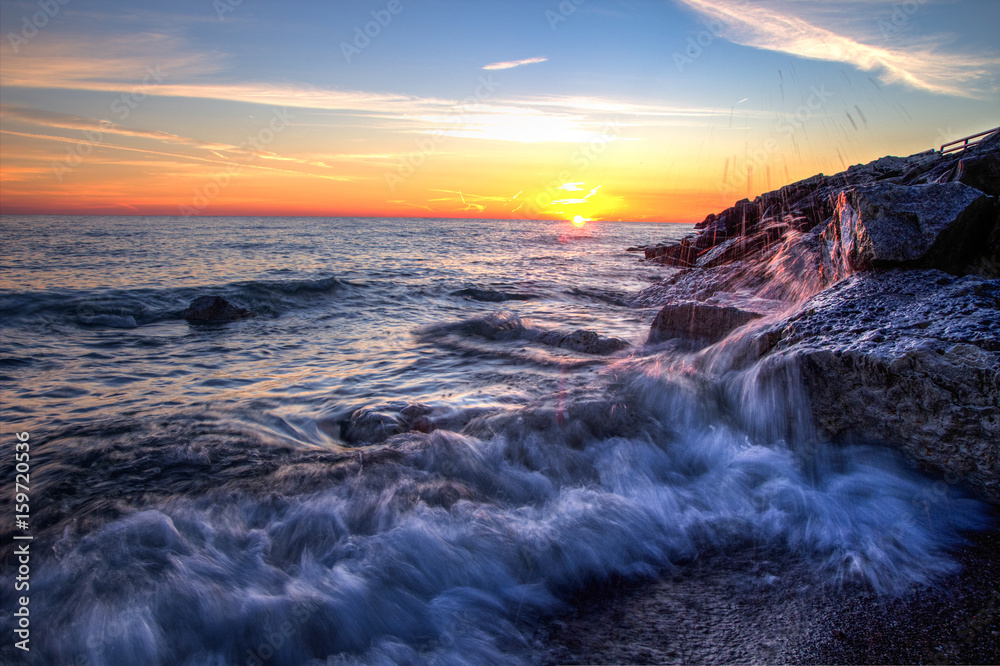 Obraz premium Beautiful Sunrise Beach Background. Waves crash onto the rocks of a Lake Huron beach with a sunrise horizon in the background. Lexington, Michigan.