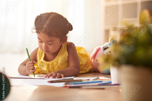 Mixed race little girl lying on floor and drawing picture with colored pencil, portrait shot photo