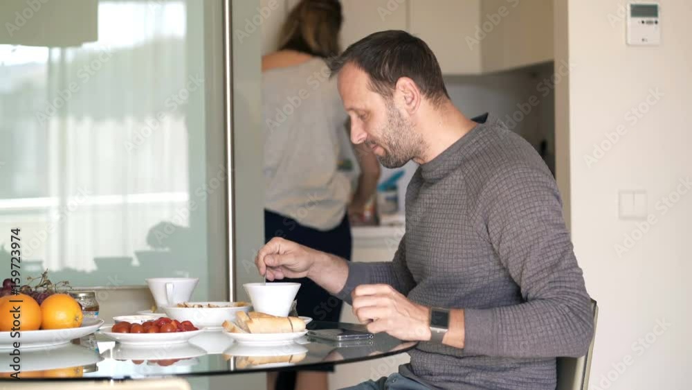 Man preparing tea during breakfast in kitchen at home, wife in the background
