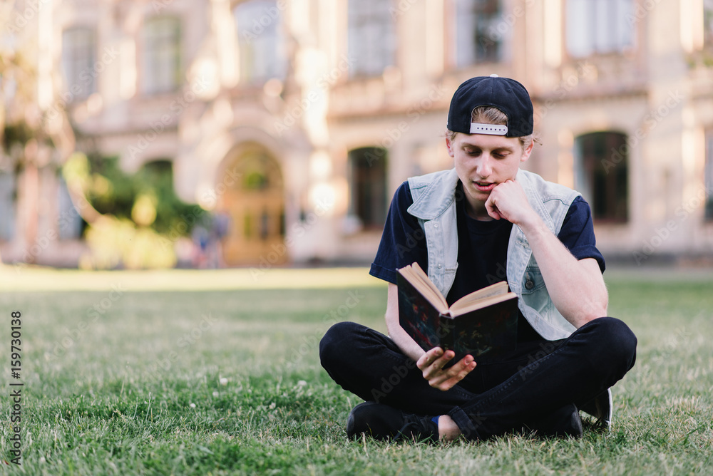 A student carefully reads a book sitting on a grass in a park near a college. Teenager reading a book outdoors Student Life