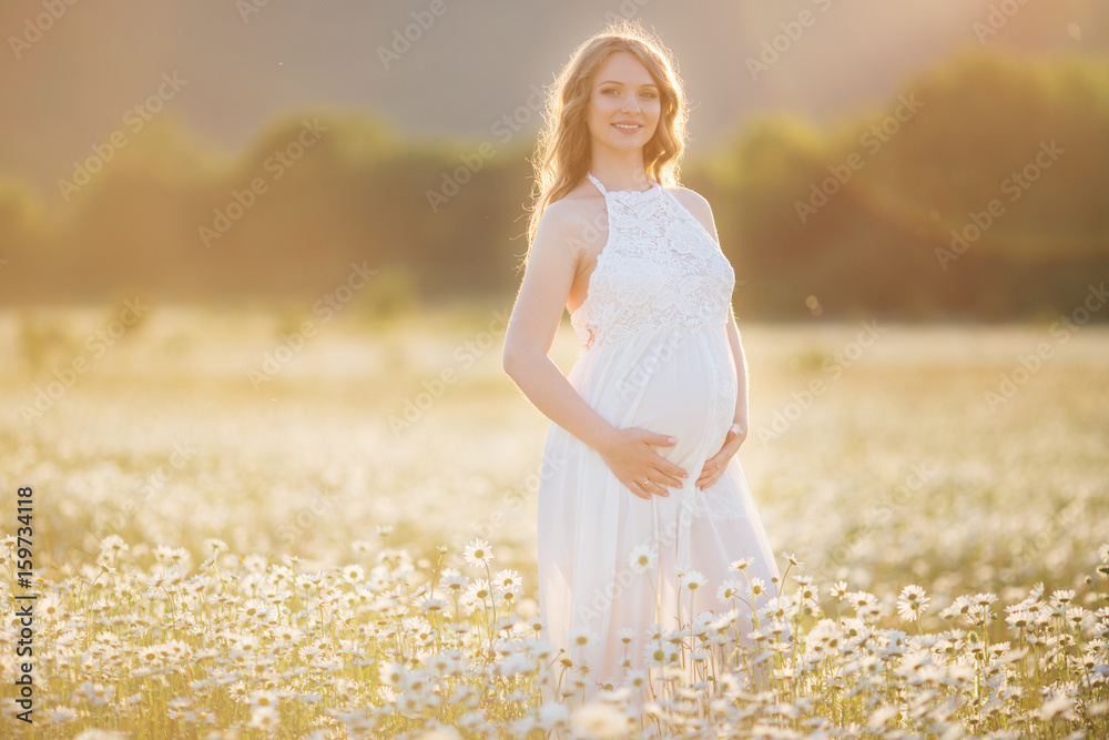 Beautiful happy pregnant girl on the field of daisy flowers in sunset lights Beautiful happy pregnant girl on the field of daisy flowers in sunset lights