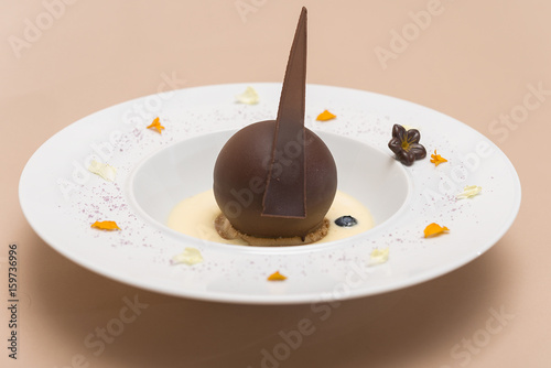 Luxurious dessert with chocolate globe and vanilla sauce, served on a biscuit, decorated with chocolate flower, flower petals and forest fruits, served in a white plate, light background, isolated