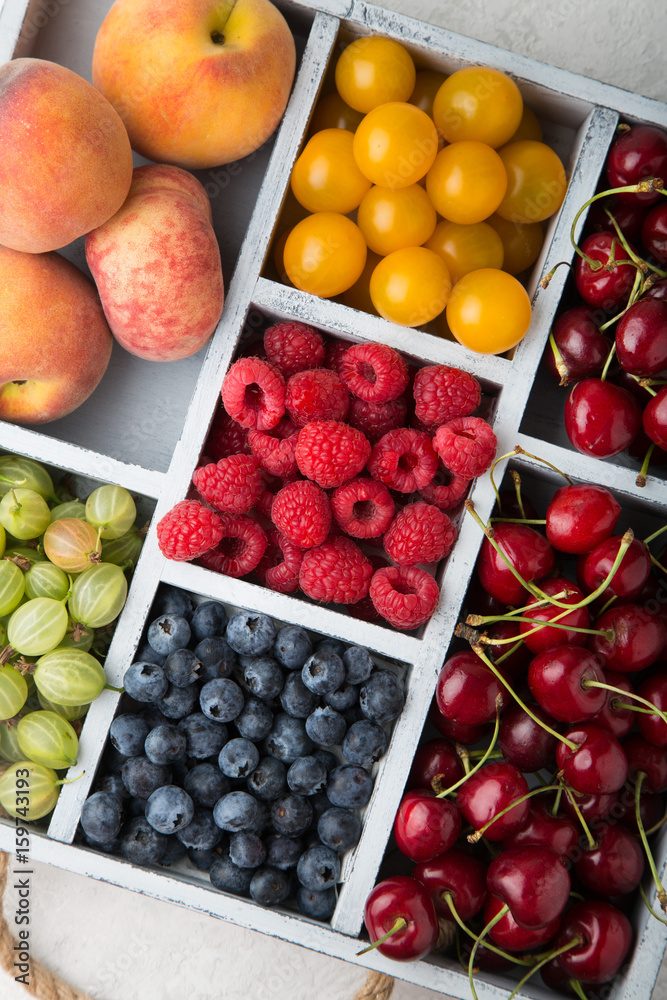 Assorted summer fruits and berries in a light wooden box with cells standing on light concrete background. Blueberries, raspberries, gooseberries, peaches, cherries. Top view