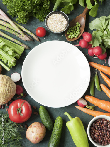 A white plate surrounded by variety of fresh raw vegetables on rustic dark green background.