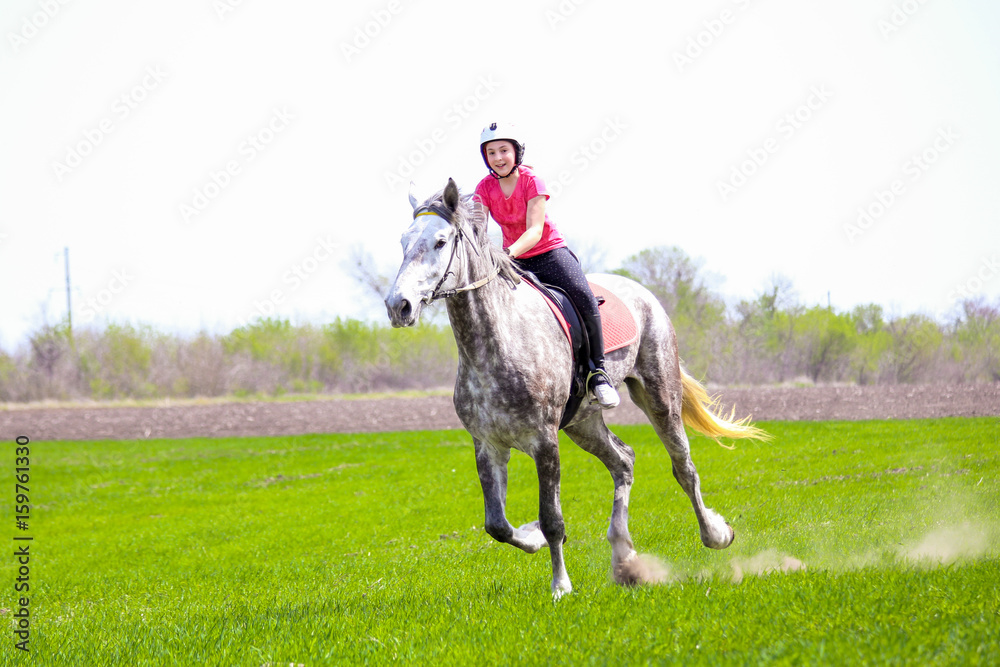 Young girl in a helmet riding a dapple-grey horse on a grass field