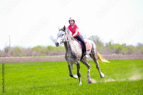 Young girl in a helmet riding a dapple-grey horse on a grass field