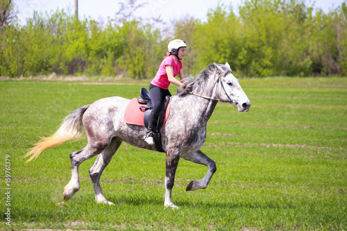 Young girl in a helmet riding a dapple-grey horse on a grass field © Kirill Gorlov