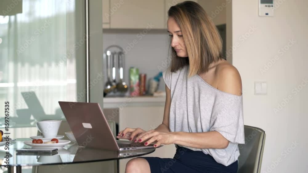 Young woman working on laptop and drinking coffee in kitchen at home
