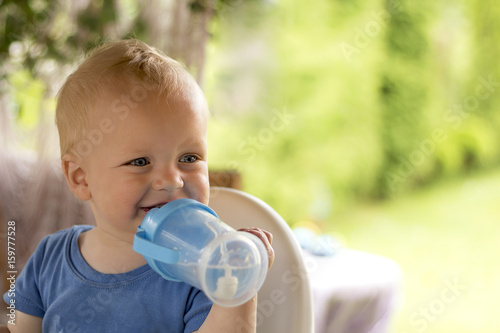 Awesome curly blondy toddler sitting outdoor and drinking water.