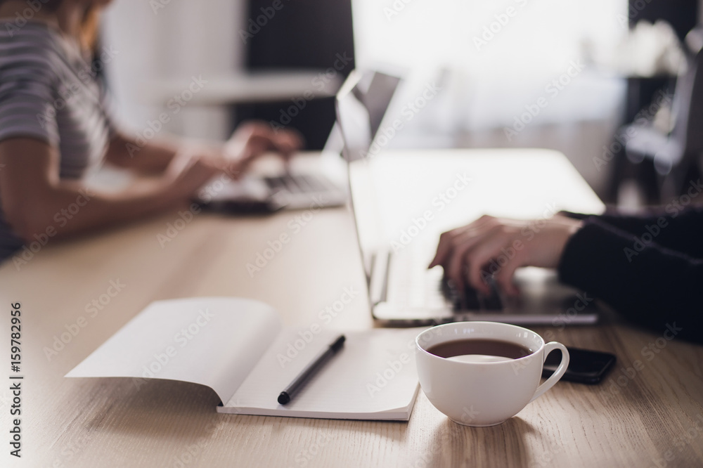 Close up picture of two businesswomen using tablet computer and laptop. Notebook with pen and cup of coffee in the foreground. Close up picture of two businesswomen using tablet computer and laptop. Notebook with pen and cup of coffee in the foreground.