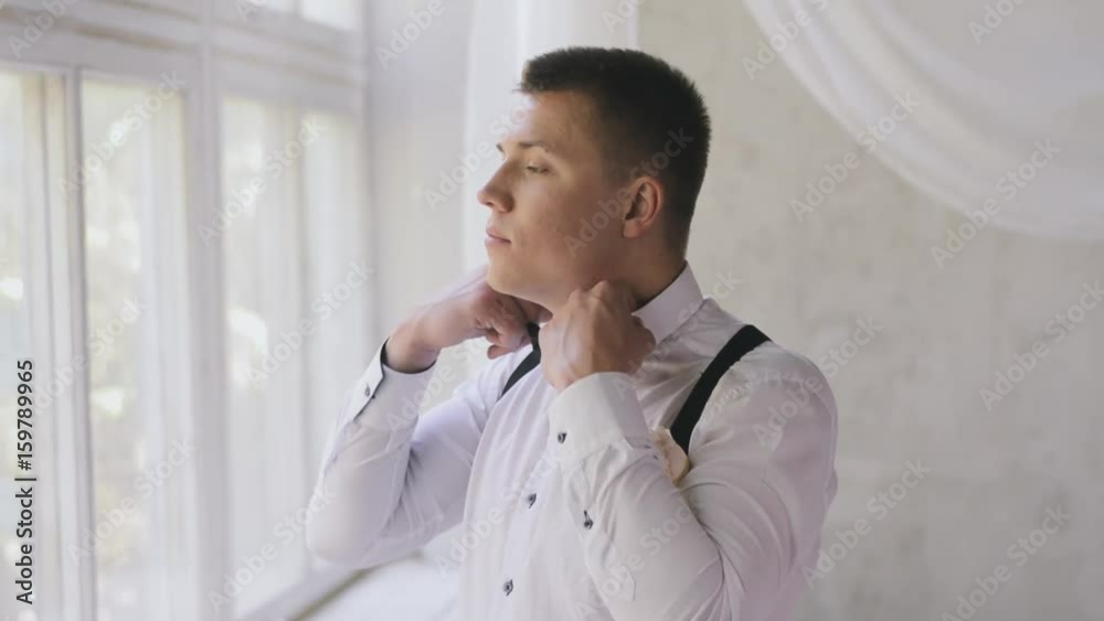 Caucasian man with a short haircut corrects a bow tie near the window in the studio. The groom in a white shirt and butterfly smiles indoors waiting for the bride. Caucasian man with a short haircut corrects a bow tie near the window in the studio. The groom in a white shirt and butterfly smiles indoors waiting for the bride.