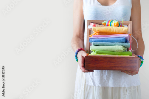 A woman holding a stack with colorful fabrics. Needle bed, scissors, measuring tape. Beige background.