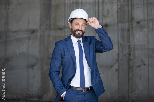 Handsome middle aged businessman in hard hat and suit standing and smiling at work