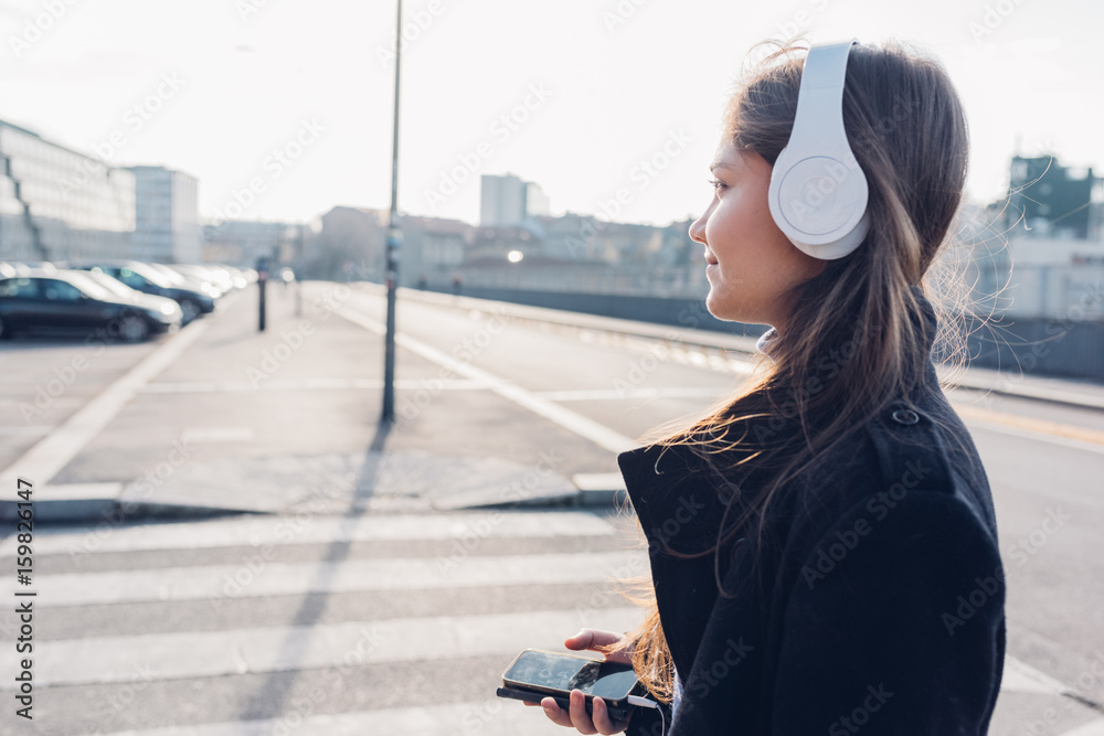 Fototapeta premium Side view young woman walking in the city back light listening music headphones with smart phone hand hold - music, relaxing, enjoyment concept