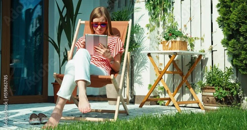 Young woman relaxing on the beach chair and using tablet in the sunny day. Young adult looking and smiling at camera