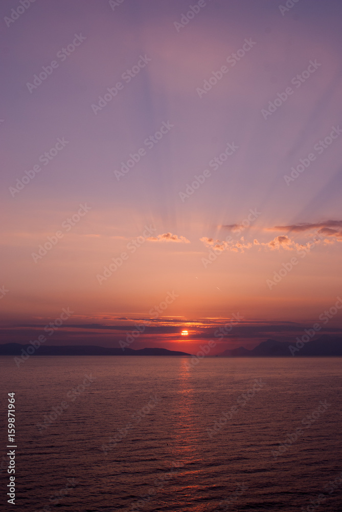 vertical image of sunset over the sea with purple blue hues and sunrays coming through clouds above
