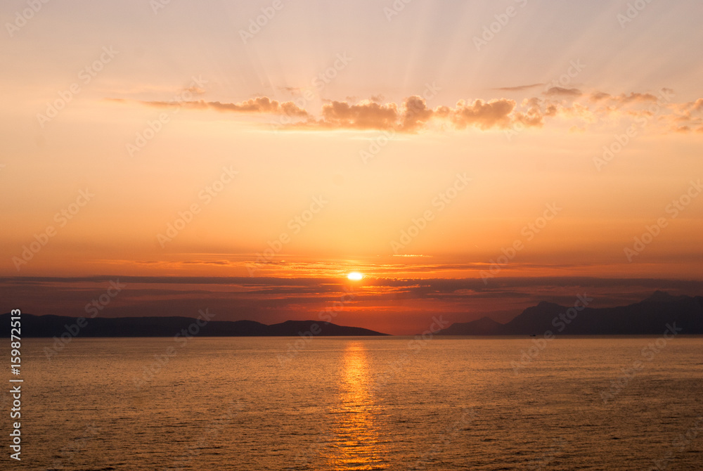 horizontal image of golden sunset with sun centrally low above the sea and sunrays coming through clouds above
