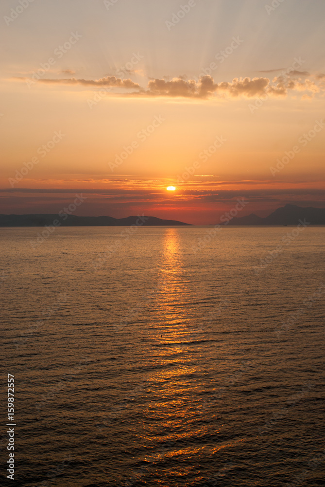 panoramic image of golden sunset with sun low above the sea and sunrays coming through clouds above
